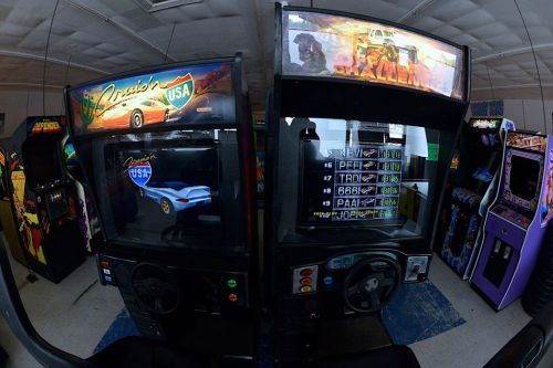 Two vintage arcade racing games side by side, featuring colorful graphics and steering wheels in a dimly lit room.