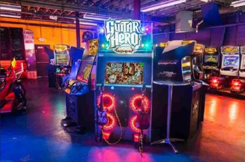 A vibrant arcade featuring a lit Guitar Hero machine surrounded by classic arcade games in a lively setting.