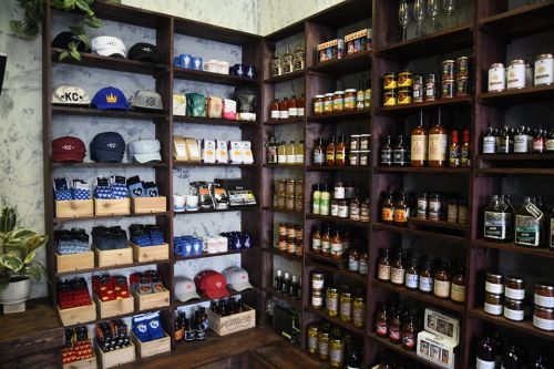 A wooden shelf filled with various products, including hats, sauces, snacks, and beverages in a cozy store setting.