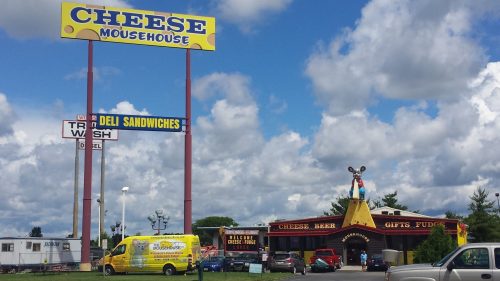 A colorful cheese-themed store with a large sign, featuring a mouse statue and parked vehicles under a cloudy sky.
