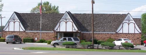 A brick building with a distinctive roof design, surrounded by greenery and parked cars, under a clear blue sky.
