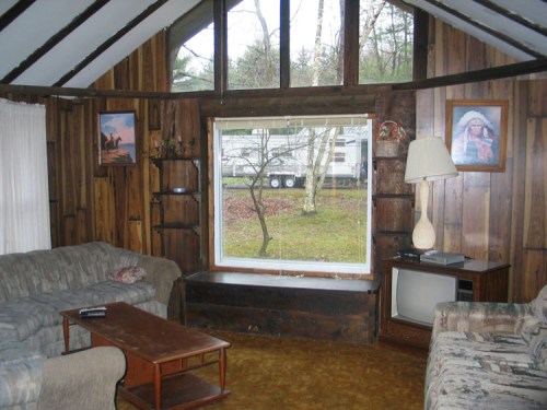 Cozy cabin interior with wooden walls, two sofas, a coffee table, a lamp, and a window overlooking a wooded area.