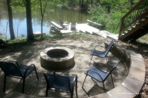 A cozy fire pit area with four chairs, surrounded by trees, overlooking a calm pond and wooden dock.
