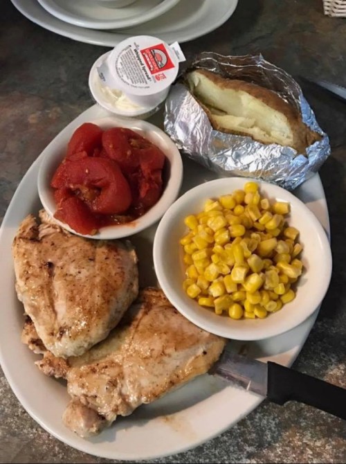 A plate with grilled chicken, baked potato, corn, and diced tomatoes, served with a small container of sauce.