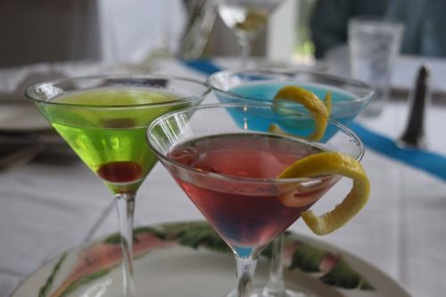Three colorful cocktails in martini glasses: green, blue, and red, garnished with lemon twists, on a table setting.
