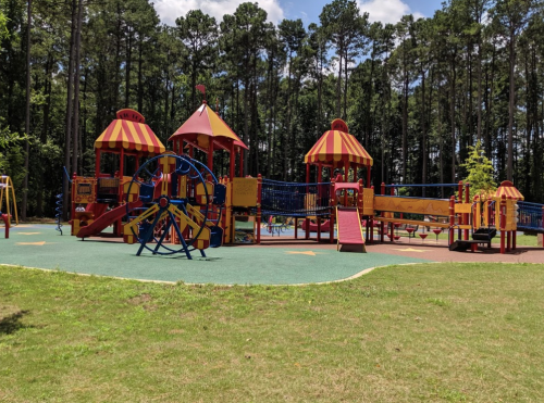Colorful playground with slides, climbing structures, and a large play area surrounded by trees and blue sky.