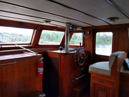 Interior of a wooden boat with a steering wheel, control panel, and large windows showing a view of water and trees.