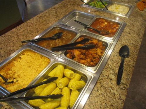 A buffet spread featuring various dishes, including macaroni and cheese, corn, chicken, and sauces in metal trays.