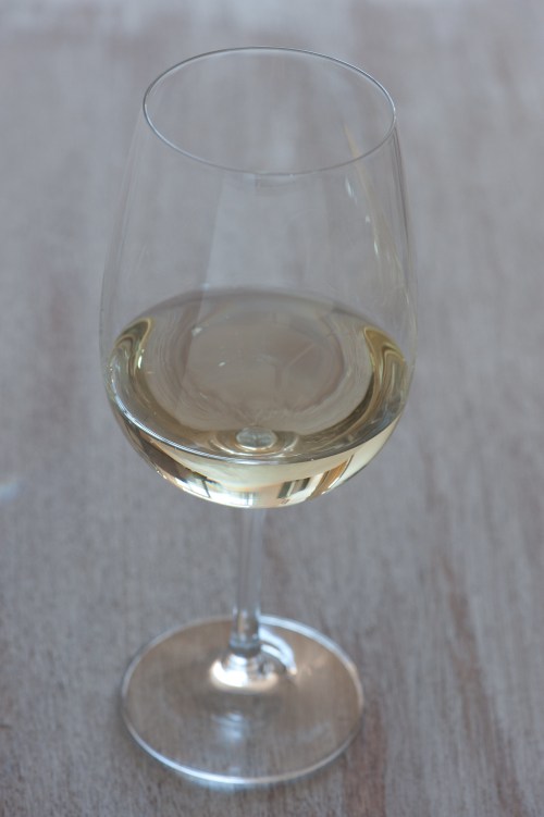 A clear wine glass filled with light-colored wine, resting on a wooden surface.