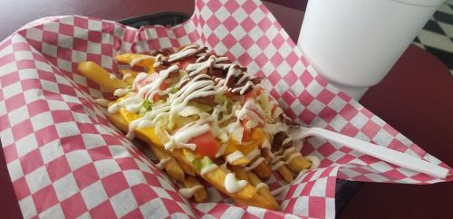 A basket of loaded fries topped with lettuce, tomato, bacon, and drizzled with mayonnaise, next to a drink.