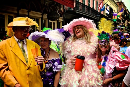 A lively group in colorful costumes and hats celebrates at a festive street event, enjoying drinks and laughter.