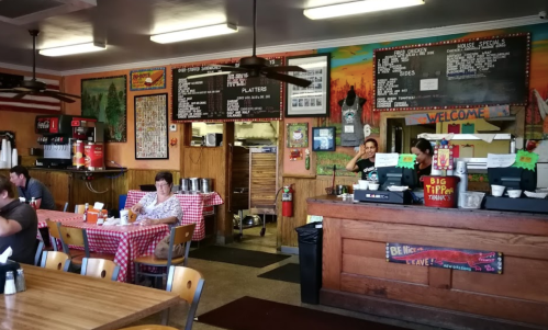 A cozy diner interior with checkered tablecloths, a counter, menu boards, and patrons enjoying their meals.