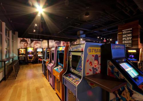 A row of colorful vintage arcade machines in a dimly lit game room with pinball machines in the background.
