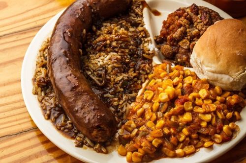 A plate of food featuring sausage, rice with gravy, corn, stuffing, and a dinner roll on a wooden table.