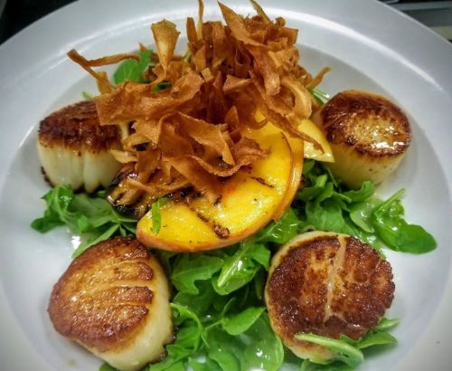 A plate of seared scallops on a bed of arugula, topped with crispy fried onions and grilled peach slices.