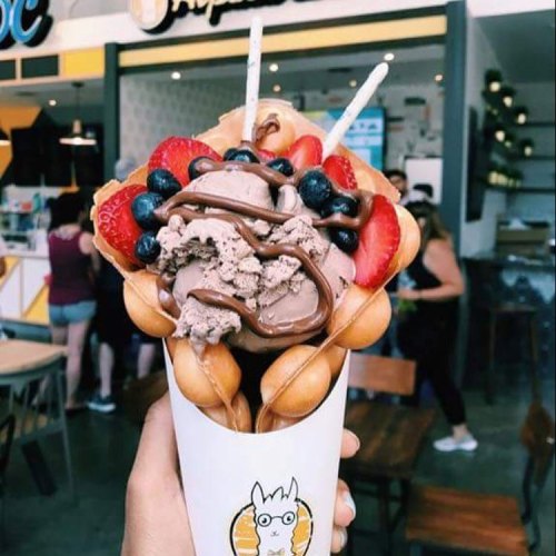 A hand holding a colorful dessert cone filled with ice cream, fruit, and drizzled chocolate, set in a vibrant café.