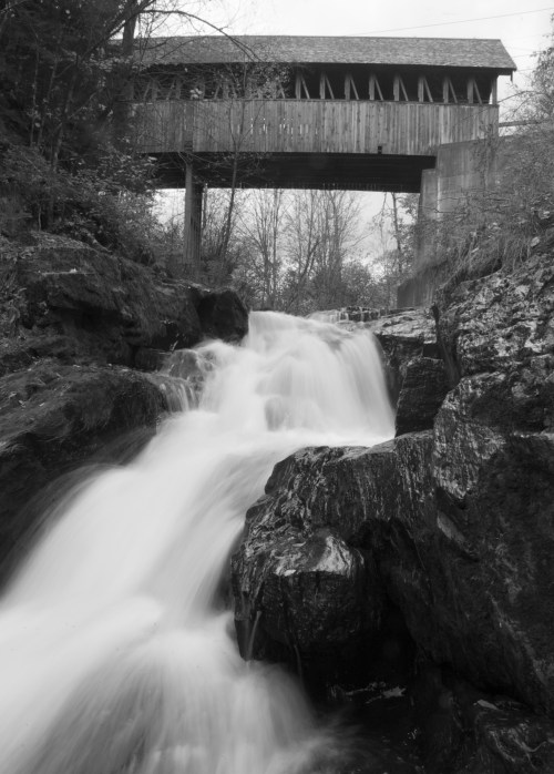 A black and white image of a waterfall cascading over rocks, with a wooden bridge above surrounded by trees.