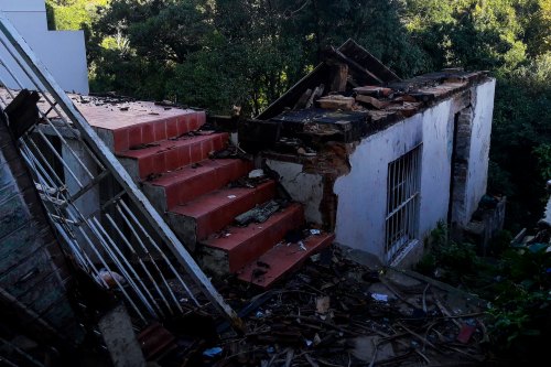 A damaged building with a collapsed roof and stairs, surrounded by overgrown vegetation and debris.
