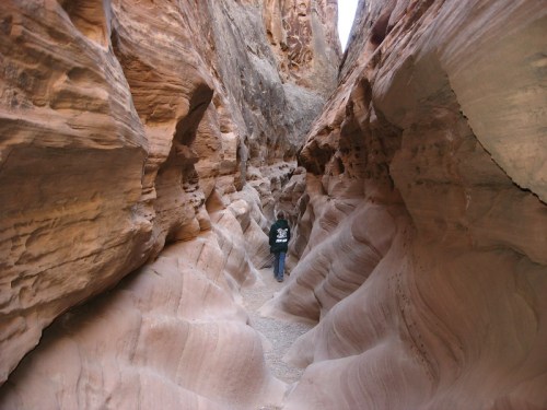 A person walks through a narrow, winding canyon with smooth, layered rock formations on either side.