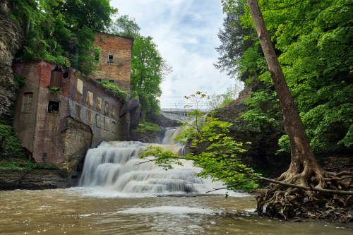 A serene waterfall cascades over rocks, surrounded by lush greenery and remnants of an old brick building.