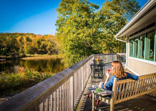 A woman relaxes on a deck by a serene lake, enjoying a drink with autumn foliage in the background.