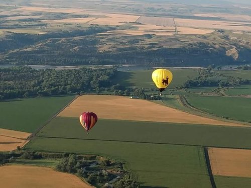 Two hot air balloons float over a patchwork of green and golden fields, with a river winding through the landscape below.