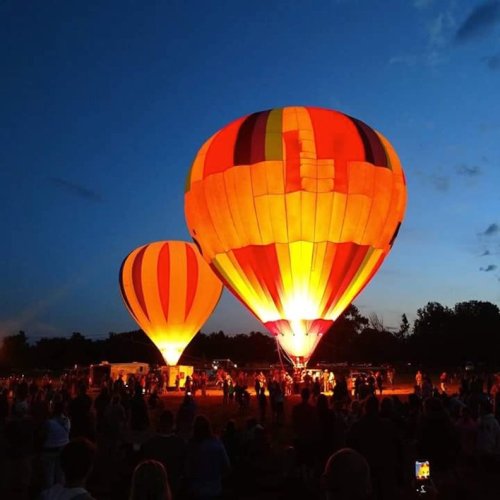 Two brightly colored hot air balloons glow against a twilight sky, surrounded by a crowd of spectators.
