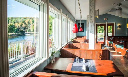 Bright restaurant interior with large windows overlooking a river, featuring wooden booths and pendant lighting.