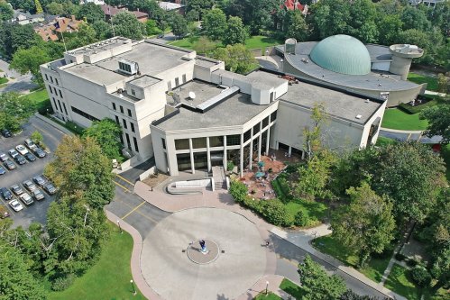 Aerial view of a modern building with a dome, surrounded by greenery and a circular plaza, with parked cars nearby.