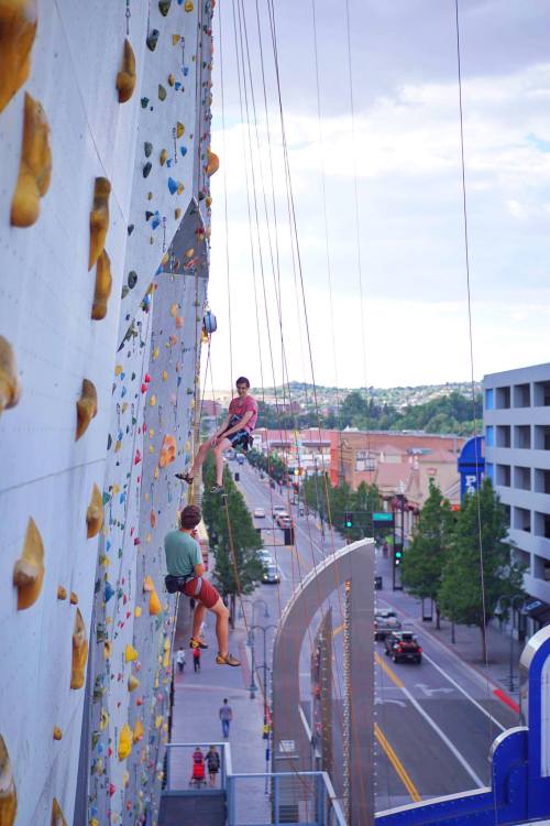 Two climbers ascend a tall rock wall in an urban setting, with buildings and trees visible below.