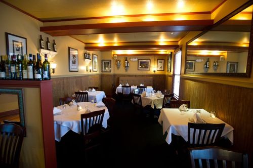 Cozy restaurant interior with white tablecloths, wooden chairs, and soft lighting, featuring wine bottles on a shelf.