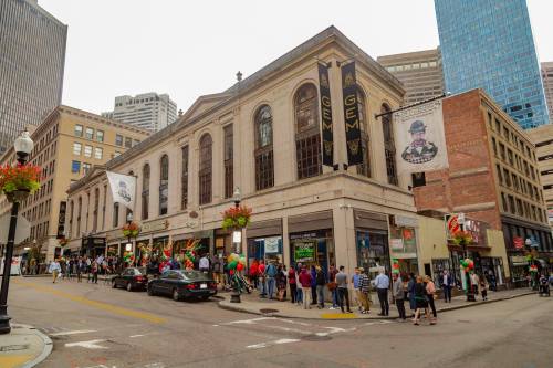 A bustling city street scene with a historic building, banners, and a long line of people outside a venue.