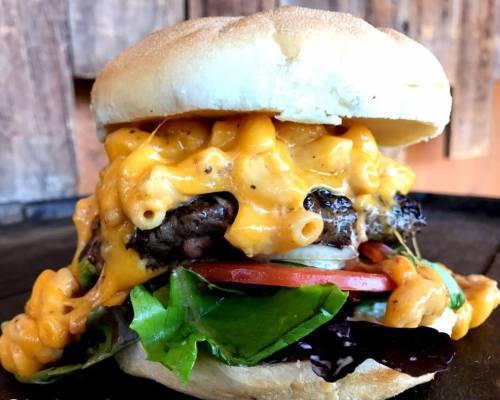 A towering burger with melted cheese, macaroni, lettuce, tomato, and a soft bun, set against a rustic wooden background.