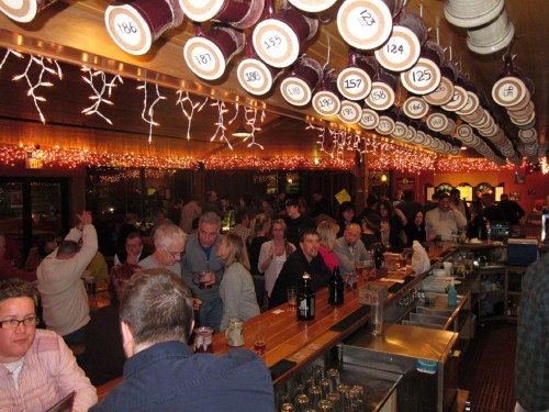 A lively bar scene with patrons socializing, string lights overhead, and a long wooden bar filled with drinks.