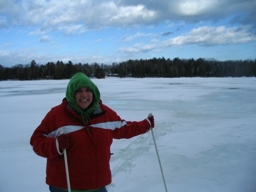 A person in a red jacket and green hood smiles while standing on a snowy landscape with a cloudy sky in the background.