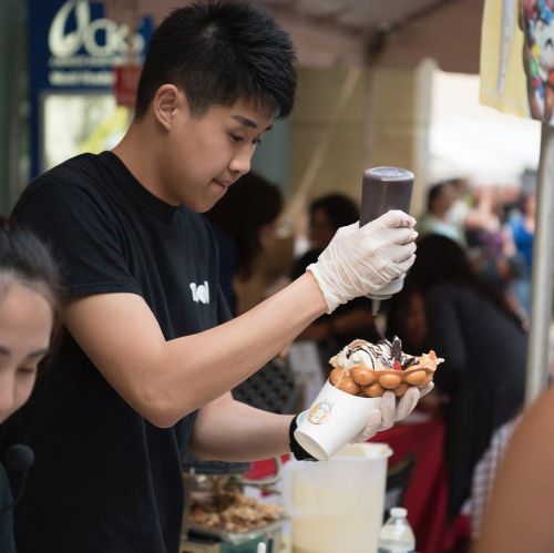 A young vendor in a black shirt carefully drizzles chocolate sauce over a dessert at a busy outdoor market.