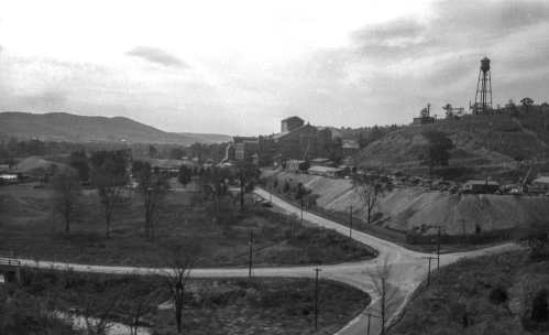 A black-and-white landscape featuring a mining town with buildings, a water tower, and winding roads amidst hills.