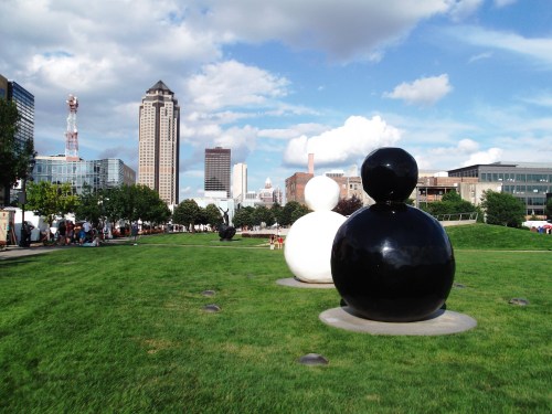 Two large, abstract sculptures in black and white on a grassy park, with city buildings and a blue sky in the background.