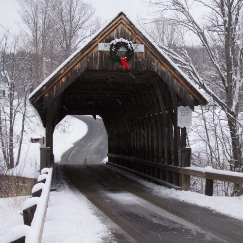 A snow-covered wooden covered bridge adorned with a wreath, leading to a winding road in a winter landscape.