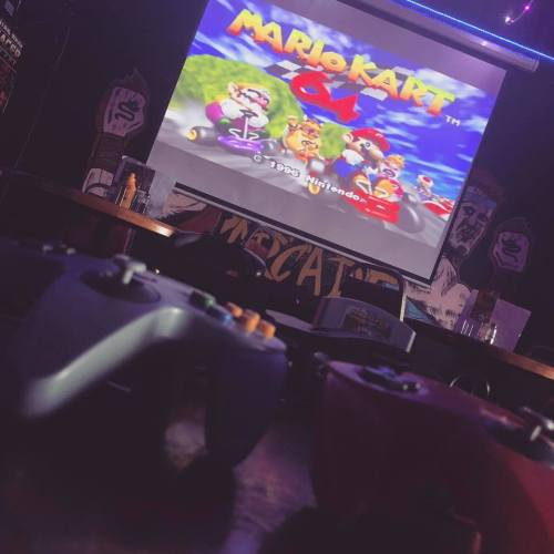 A close-up of game controllers in front of a screen displaying Mario Kart 64 in a dimly lit gaming venue.