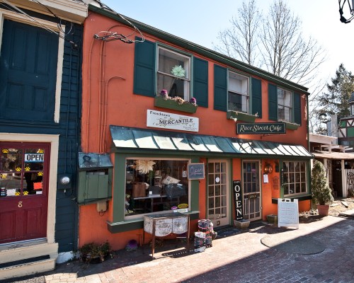Charming storefronts of a mercantile and café with colorful facades and open signs in a quaint town setting.