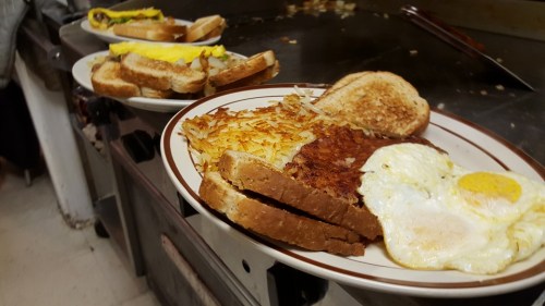 Plates of breakfast featuring eggs, toast, hash browns, and an omelet on a griddle in a diner setting.