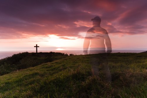 A silhouette of a person stands on a grassy hill, with a cross and a colorful sunset over the ocean in the background.