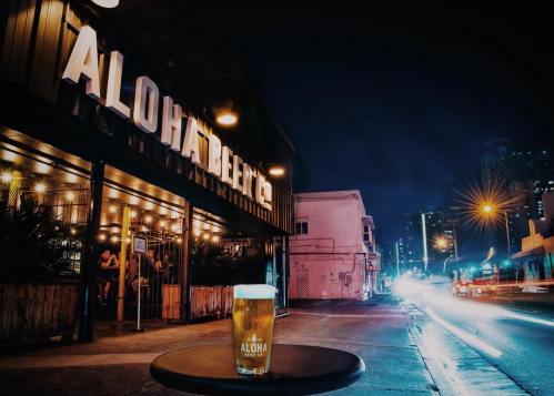 A glass of beer sits on a table outside Aloha Beer Co. at night, with city lights and a lively atmosphere in the background.