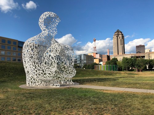 A large, white sculpture made of letters sits in a grassy area with buildings and a blue sky in the background.