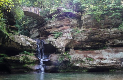 A serene waterfall cascades over rocky cliffs, surrounded by lush greenery and an old stone bridge above.