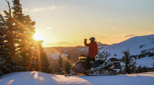 A person in a red jacket stands on a snowmobile, watching the sunset over a snowy mountain landscape.