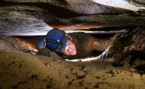 A person in a helmet crawls through a narrow cave passage, surrounded by rock and sand, with light illuminating the space.
