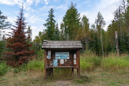 A wooden information kiosk surrounded by trees and grass, featuring a scenic image and various informational panels.