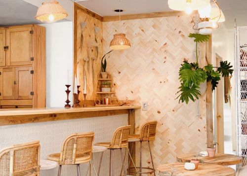 Cozy interior featuring a wooden bar, rattan stools, and a textured wall with plants and warm lighting.